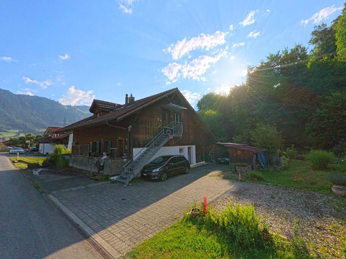In einem Wohnquartier von Zwieselberg (PLZ 3647, Kanton Bern) steht ein dunkles Haus mit Aussentreppe und angebautem Carport am Waldrand, waehrend die Sonne hinter den Baeumen hervorstrahlt.